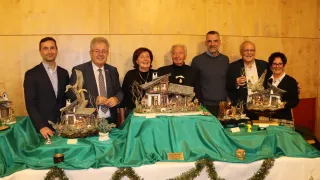 Eine Gruppe von Personen, feierlich gekleidet, posiert für ein Foto vor einem geschmückten Tisch mit Krippenszenen, einer Kulisse aus Holzpaneelen und festlichen Dekorationen.