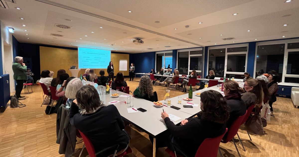Eine Gruppe von Frauen sitzt in einem Konferenzraum um einen langen Tisch und hört einer Präsentation zu. Eine Frau steht vor einer Leinwand und eine andere neben einer Tafel.