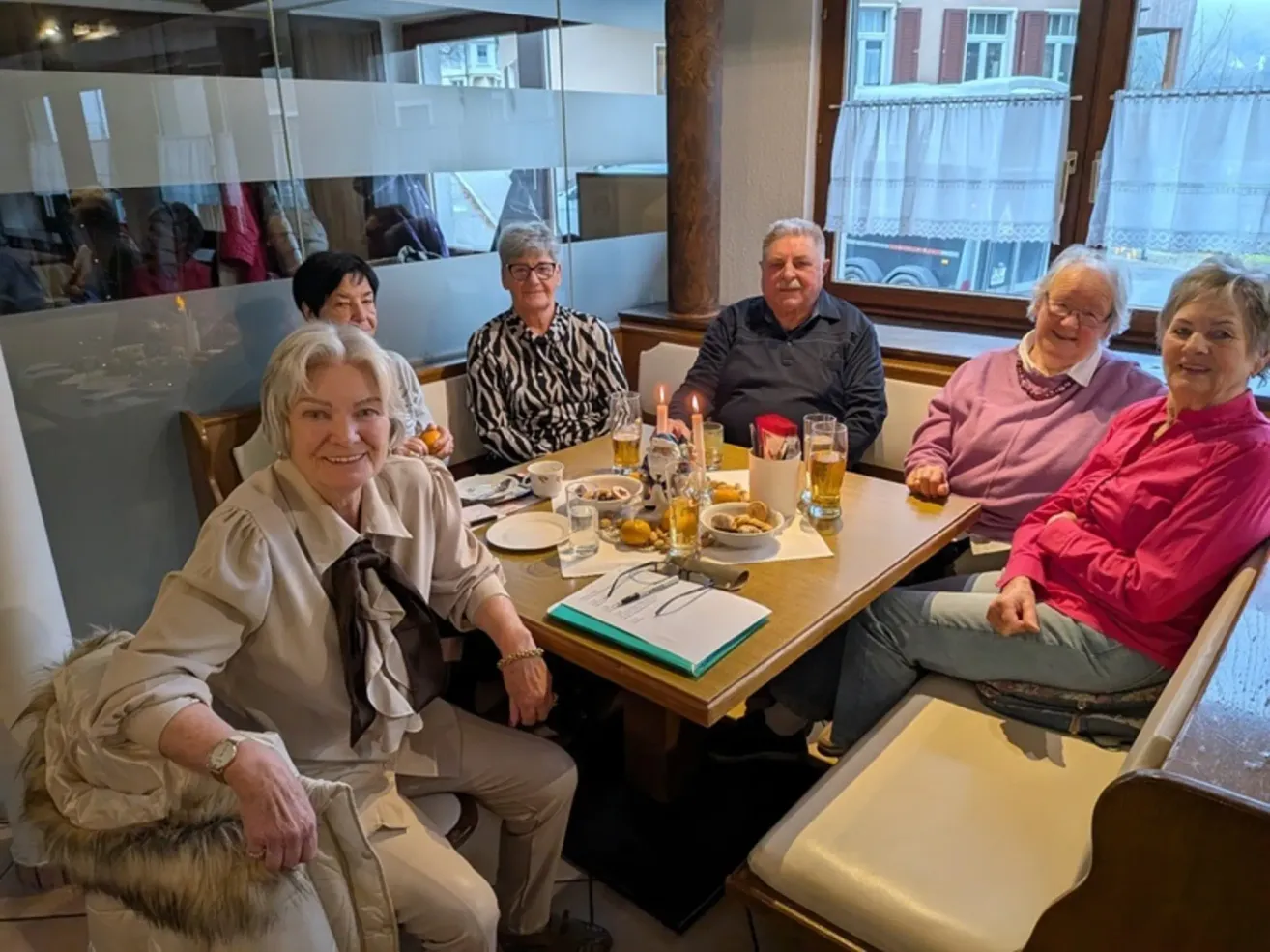 Eine Gruppe älterer Menschen versammelt sich in einem Restaurant. Sie sitzen um einen Tisch herum mit Essen, Getränken und Kerzen. Sie lächeln und scheinen ihre Mahlzeit zu genießen.
