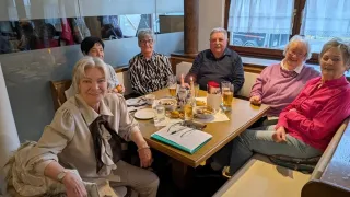 Eine Gruppe älterer Menschen versammelt sich in einem Restaurant. Sie sitzen um einen Tisch herum mit Essen, Getränken und Kerzen. Sie lächeln und scheinen ihre Mahlzeit zu genießen.