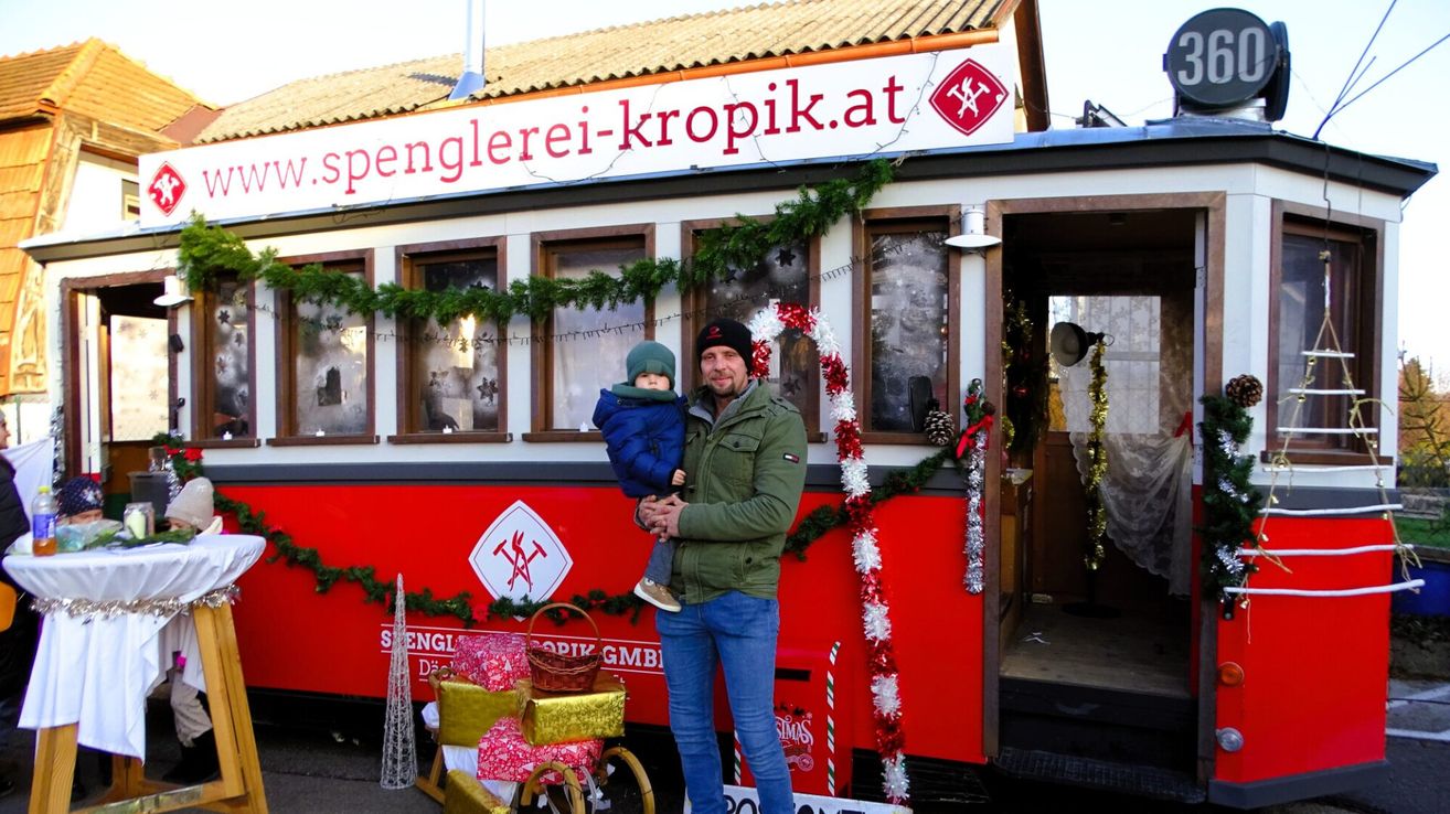 Ein Mann hält ein Kind vor einem dekorierten Straßenbahnwagen mit der Aufschrift 'spenglerei-kropik.at'. Sie sind beide warm gekleidet, und der Wagen ist mit Weihnachtsdekoration geschmückt.