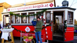 Ein Mann hält ein Kind vor einem dekorierten Straßenbahnwagen mit der Aufschrift 'spenglerei-kropik.at'. Sie sind beide warm gekleidet, und der Wagen ist mit Weihnachtsdekoration geschmückt.