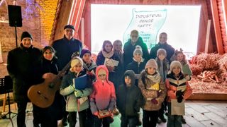 Eine Gruppe von Kindern und Erwachsenen steht vor einem Banner mit der Aufschrift 'Industrial Dance Party'. Die Kinder tragen Winterkleidung und einige halten Urkunden. Zwei Männer, einer mit einer Gitarre, stehen hinter ihnen.