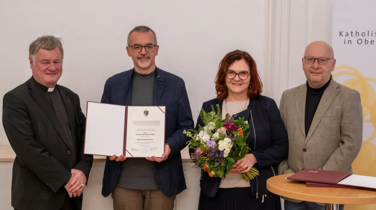 A man and a woman stand next to each other holding a certificate and a bouquet of flowers. The man is wearing glasses and a blue blazer, and the woman is wearing a blue jacket and holding a bouquet. Behind them is a white wall.