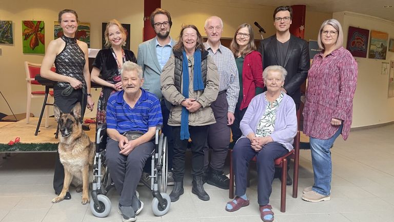 Eine Gruppe von Menschen, einige sitzend und andere stehend, posiert für ein Foto. Eine Frau im Rollstuhl ist in der Mitte. Sie alle sehen glücklich aus.