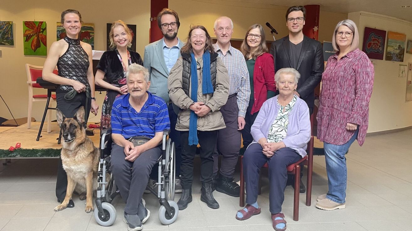 Eine Gruppe von Menschen, einige sitzend und andere stehend, posiert für ein Foto. Eine Frau im Rollstuhl ist in der Mitte. Sie alle sehen glücklich aus.