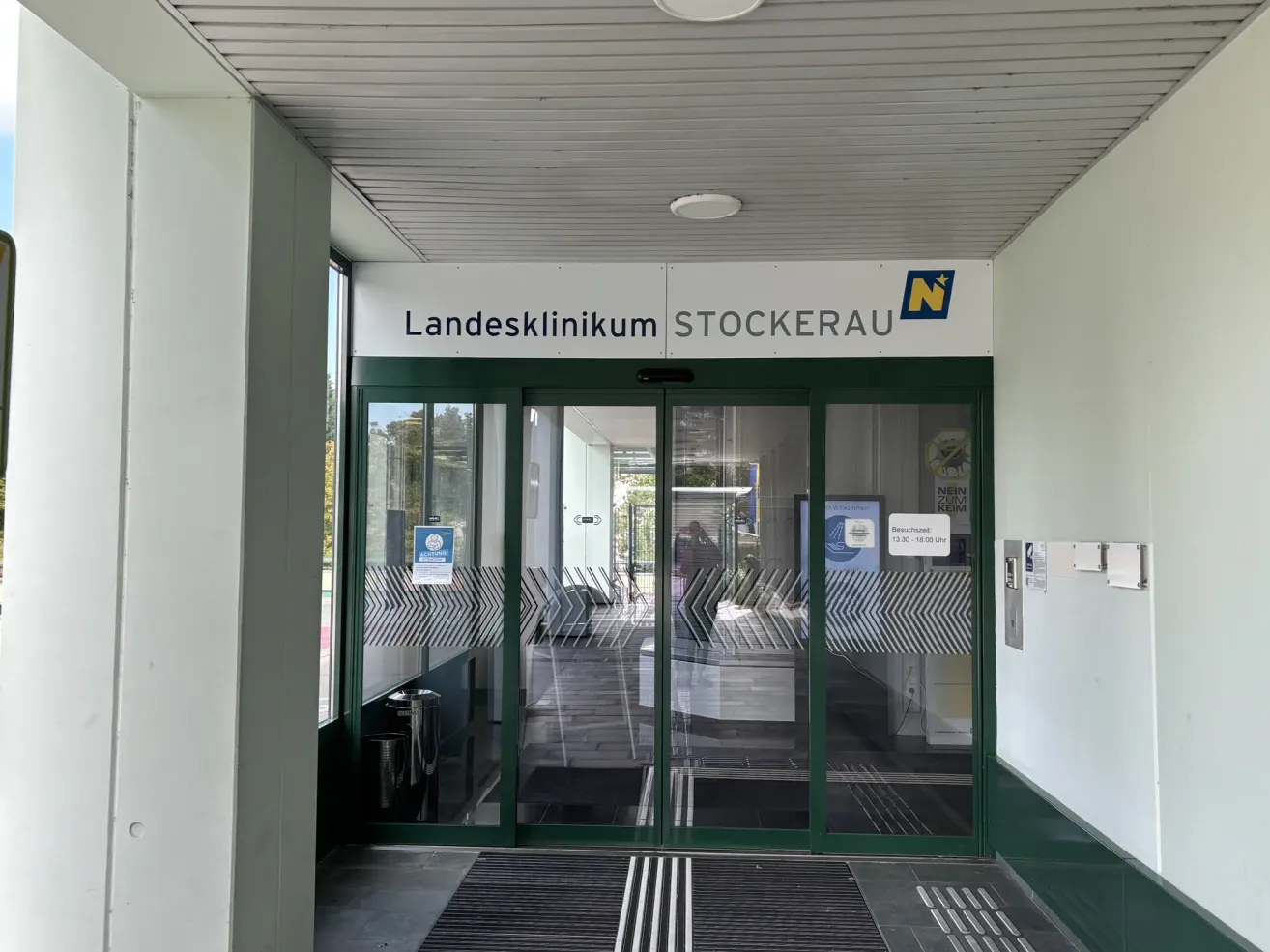 Der Eingang des Landesklinikums Stockerau. Es zeigt Glastüren, ein reflektierendes Schild und eine weiße Wand mit Beschilderung.