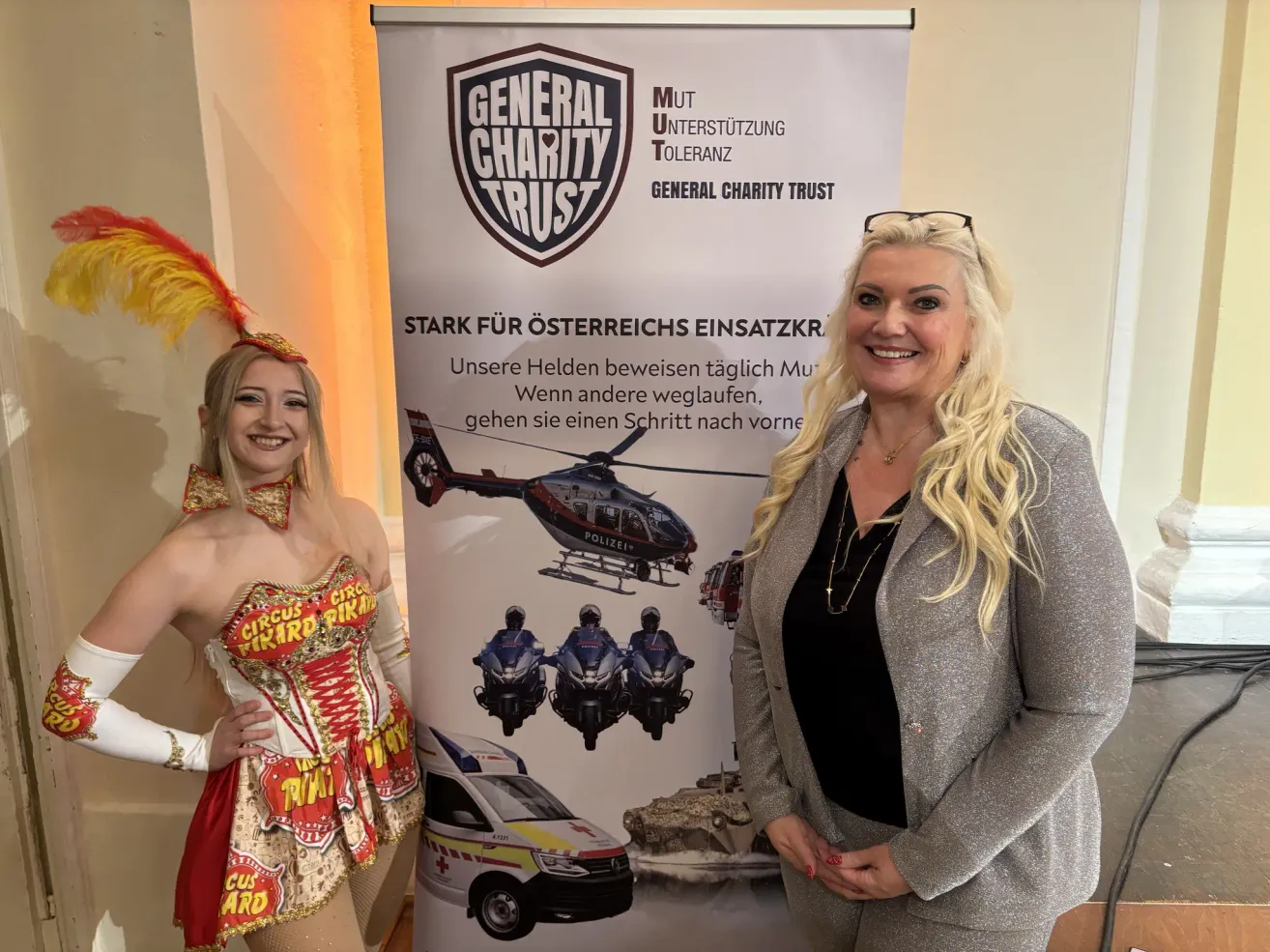 Zwei Frauen stehen neben einem Banner mit dem Logo des General Charity Trust, einem Hubschrauber und verschiedenen Rettungsfahrzeugen. Eine Frau trägt ein Kostüm.