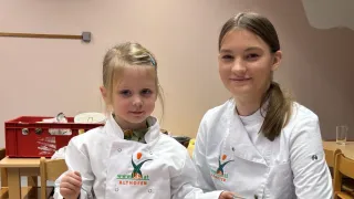 Zwei Mädchen in weißen Kochjacken in einer Küche, eine hält einen Spatel, stehen neben einem Tisch mit Schüsseln, und ein roter Kasten dahinter.