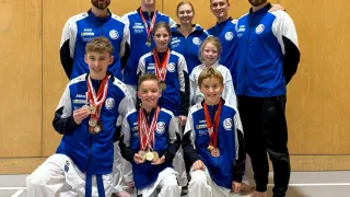 Eine Gruppe junger Athleten, sowohl männlich als auch weiblich, in blauen und weißen Uniformen und mit Medaillen, posiert für ein Foto in einer Turnhalle. Sie stehen und knien auf einer blauen Linie auf dem Boden.