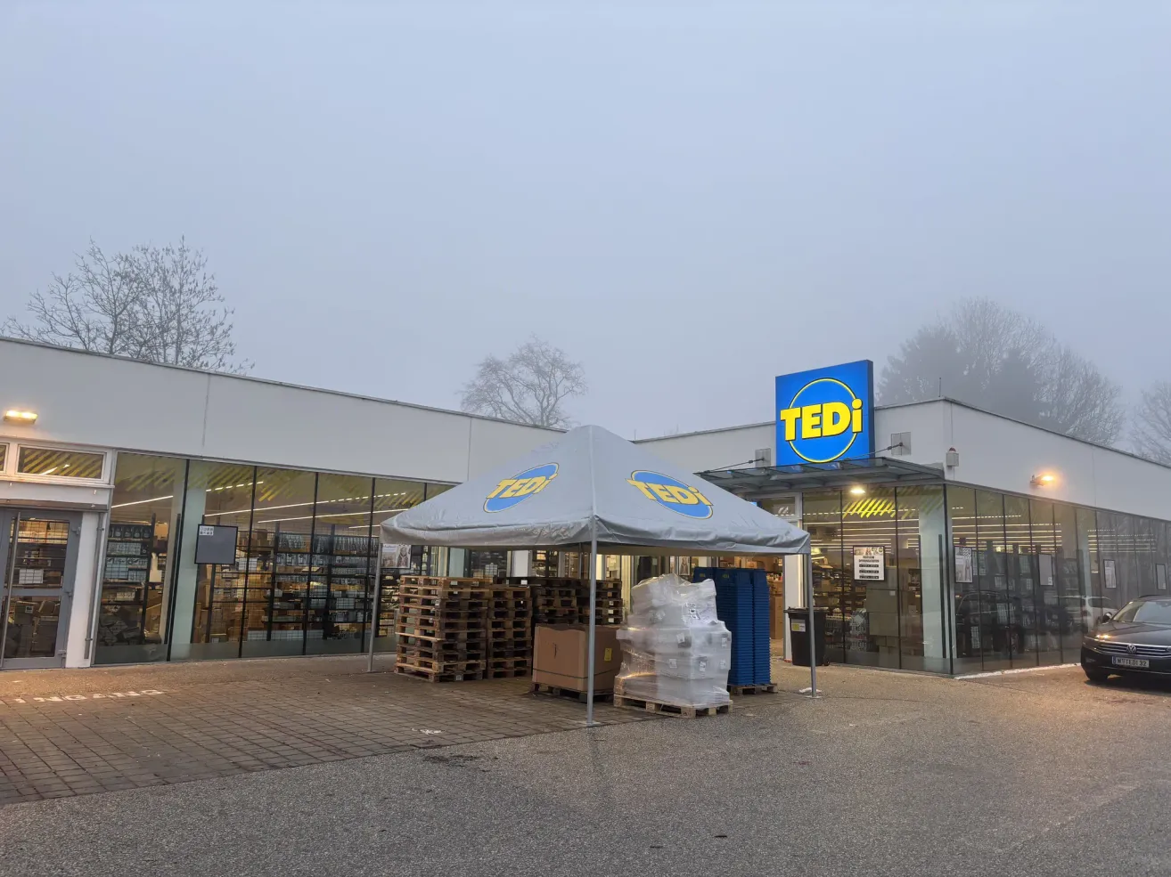 Außenansicht eines Tedi-Geschäfts an einem nebligen Tag mit einem Zelt und gestapelten Kisten davor.