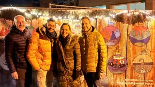 Vier Personen in Winterkleidung posieren vor einer dekorierten Holzwand, möglicherweise bei einer Veranstaltung. Die Wand ist mit Ornamenten und Lichtern geschmückt und es befindet sich ein Wasserzeichen mit der Aufschrift 'druck at' in der Ecke.