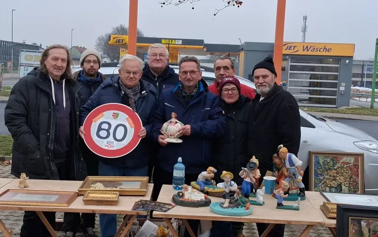 Eine Gruppe von Menschen steht hinter einem Tisch, auf dem verschiedene Figuren und Kunstwerke ausgestellt sind. Ein Mann auf der linken Seite hält ein Schild mit der Nummer 80.