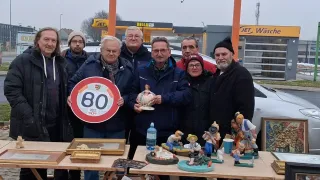 Eine Gruppe von Menschen steht hinter einem Tisch, auf dem verschiedene Figuren und Kunstwerke ausgestellt sind. Ein Mann auf der linken Seite hält ein Schild mit der Nummer 80.