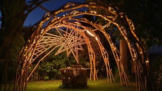 Ein hölzerne Laube mit sternförmigem Design ist bei Nacht mit warmen Lichtern beleuchtet. Sie verfügt über einen zentralen offenen Bereich mit einem Holztisch und ist von Grün umgeben.
