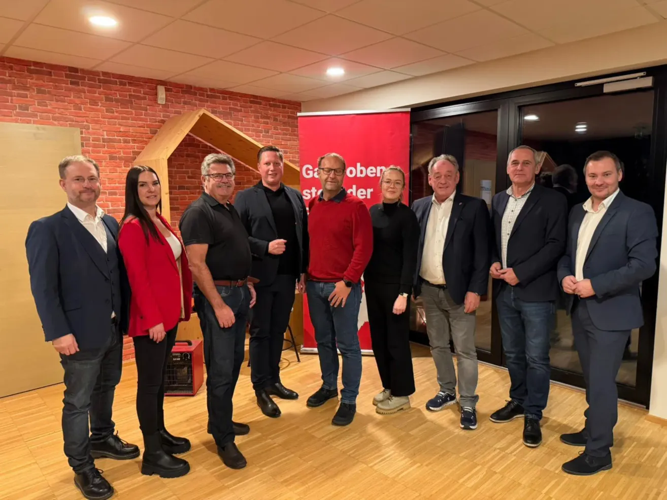 Neun Personen, sieben Männer und zwei Frauen, stehen in einer Reihe in einem Raum. Sie sind schick gekleidet. Ein roter Banner mit Text steht hinter ihnen. Eine Holzstruktur befindet sich auf der linken Seite.