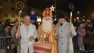 Zwei Männer in Anzügen stehen neben einem Mann in einer Sankt-Nikolaus-Kostüm, der einen Korb und Glocken trägt. Sie sind draußen bei Nacht mit festlichen Dekorationen im Hintergrund.