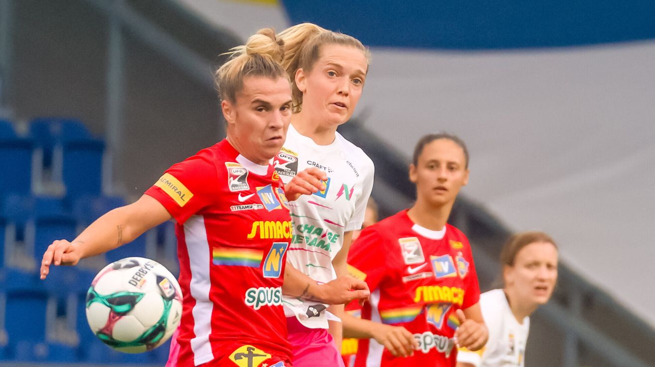 Drei Athletinnen in roten Trikots spielen Fußball, eine mit einem Ball. Die Spielerin in der Mitte schaut nach vorne, während die anderen etwas anschauen.