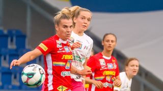 Drei Athletinnen in roten Trikots spielen Fußball, eine mit einem Ball. Die Spielerin in der Mitte schaut nach vorne, während die anderen etwas anschauen.