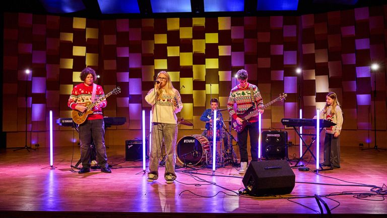 Eine Band tritt mit verschiedenen Instrumenten auf der Bühne auf. Die Sängerin hält ein Mikrofon, während die Gitarristen ihre Gitarren spielen. Der Schlagzeuger sitzt hinter dem Schlagzeug. Die Bühne ist mit einem farbenfrohen Hintergrund beleuchtet.