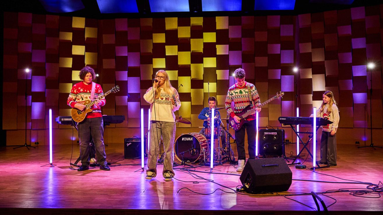 Eine Band tritt mit verschiedenen Instrumenten auf der Bühne auf. Die Sängerin hält ein Mikrofon, während die Gitarristen ihre Gitarren spielen. Der Schlagzeuger sitzt hinter dem Schlagzeug. Die Bühne ist mit einem farbenfrohen Hintergrund beleuchtet.