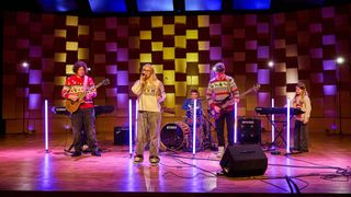 Eine Band tritt mit verschiedenen Instrumenten auf der Bühne auf. Die Sängerin hält ein Mikrofon, während die Gitarristen ihre Gitarren spielen. Der Schlagzeuger sitzt hinter dem Schlagzeug. Die Bühne ist mit einem farbenfrohen Hintergrund beleuchtet.