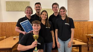 Eine Gruppe von sechs Personen in schwarzen Hemden, wahrscheinlich bei einer Schulveranstaltung, posiert für ein Foto, wobei eine Person einen Pokal hält.