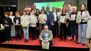 Eine Gruppe von Menschen auf einer Bühne hält Urkunden, ein Mann sitzt vorne und hält eine Urkunde. Dahinter sind zwei Banner angebracht, eines mit der Aufschrift 'Kleine' und das andere mit 'Lin'.