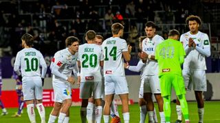 Eine Fußballmannschaft versammelt sich auf dem Feld, wobei die Spieler weiße Trikots tragen, eines davon grün. Sie scheinen eine Strategie zu besprechen.