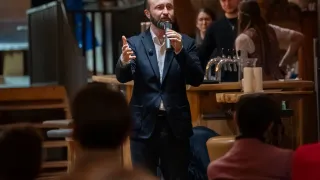 Ein Mann in einem Anzug steht vor einer Menschenmenge und spricht in ein Mikrofon. Hinter ihm stehen zwei Personen an einer Bartheke. Die Umgebung scheint ein soziales Ereignis zu sein.