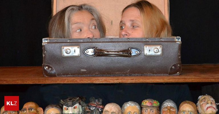 Zwei Frauen, die aus einem alten Koffer herausschauen, darunter verschiedene bemalte Holzgesichter.