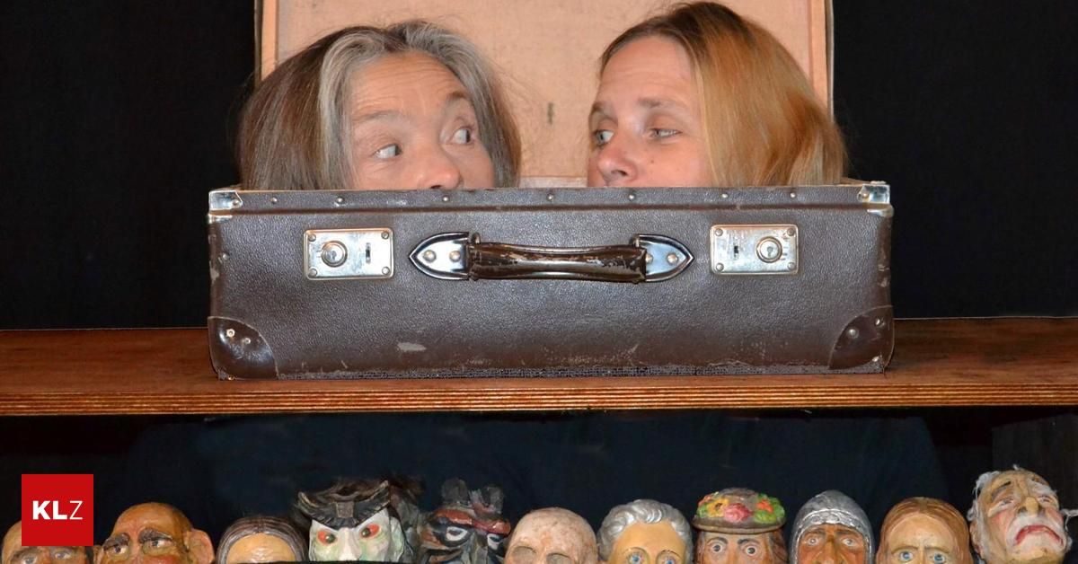 Zwei Frauen, die aus einem alten Koffer herausschauen, darunter verschiedene bemalte Holzgesichter.