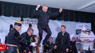 Eine Gruppe von Männern in Uniform auf der Bühne, einer springt in der Luft, andere stehen und spielen Instrumente, mit einem Banner im Hintergrund.