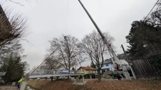 Ein Kran hebt eine Brücke über einen Kanal. Arbeiter stehen in der Nähe, und ein blaues Banner hängt an der Brücke. Bäume umgeben das Gebiet, und Gebäude sind im Hintergrund sichtbar.