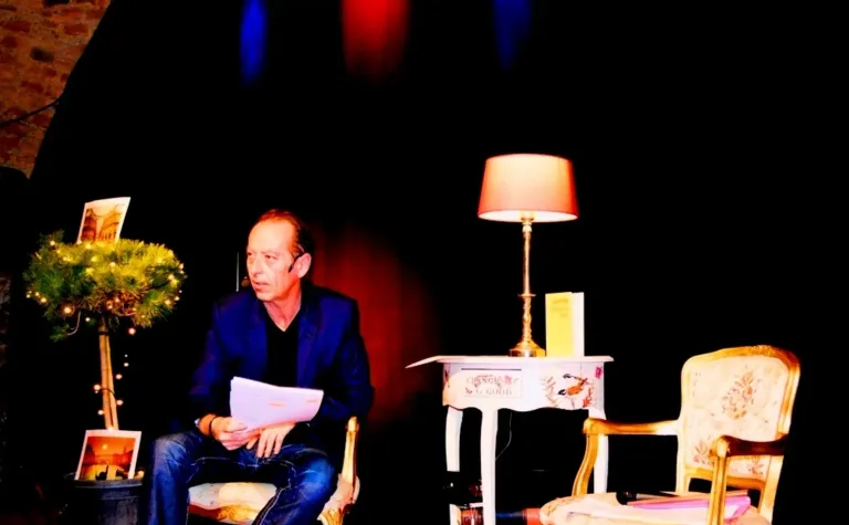 Ein Mann sitzt auf einem Stuhl, hält ein Papier in der Hand und scheint es zu lesen. Ein Lampenschirm und ein Tisch mit einem Buch stehen neben ihm.