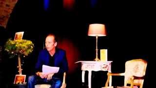 Ein Mann sitzt auf einem Stuhl, hält ein Papier in der Hand und scheint es zu lesen. Ein Lampenschirm und ein Tisch mit einem Buch stehen neben ihm.