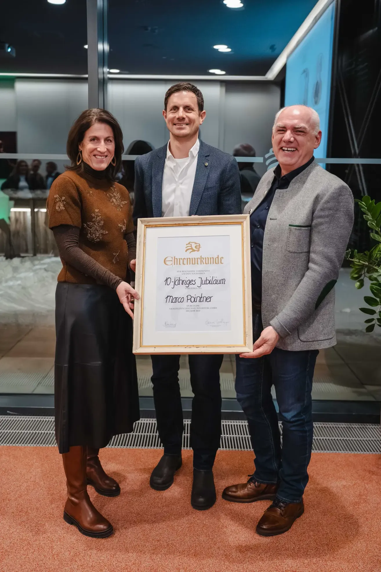 Drei Personen halten ein gerahmtes Zertifikat vor einer Glaswand und lächeln. Die Frau links trägt ein braunes Oberteil und einen schwarzen Rock. Der Mann in der Mitte trägt einen blauen Mantel und ein weißes Hemd. Der Mann rechts trägt einen grauen Mantel und blaue Jeans.