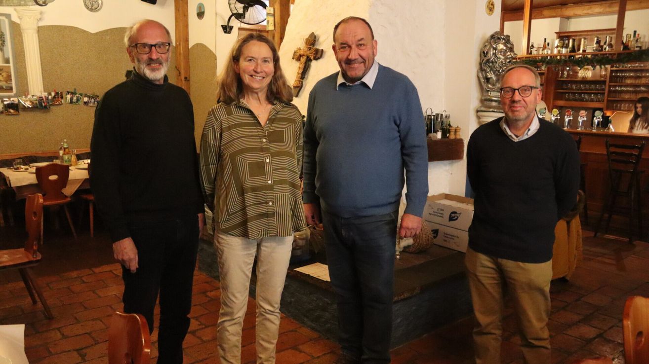 Vier Menschen stehen in einem Raum mit Ziegelboden. Sie lächeln und posieren für ein Foto. Die Frau trägt ein langärmeliges Hemd und Hose. Der Mann in der Mitte trägt einen Pullover und Jeans. Hinter ihnen steht ein Regal mit einem Kreuz und einer Statue.