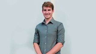 Ein Mann in einem grauen Hemd lächelt und posiert für ein Foto. Seine Hände sind vor ihm verschränkt.