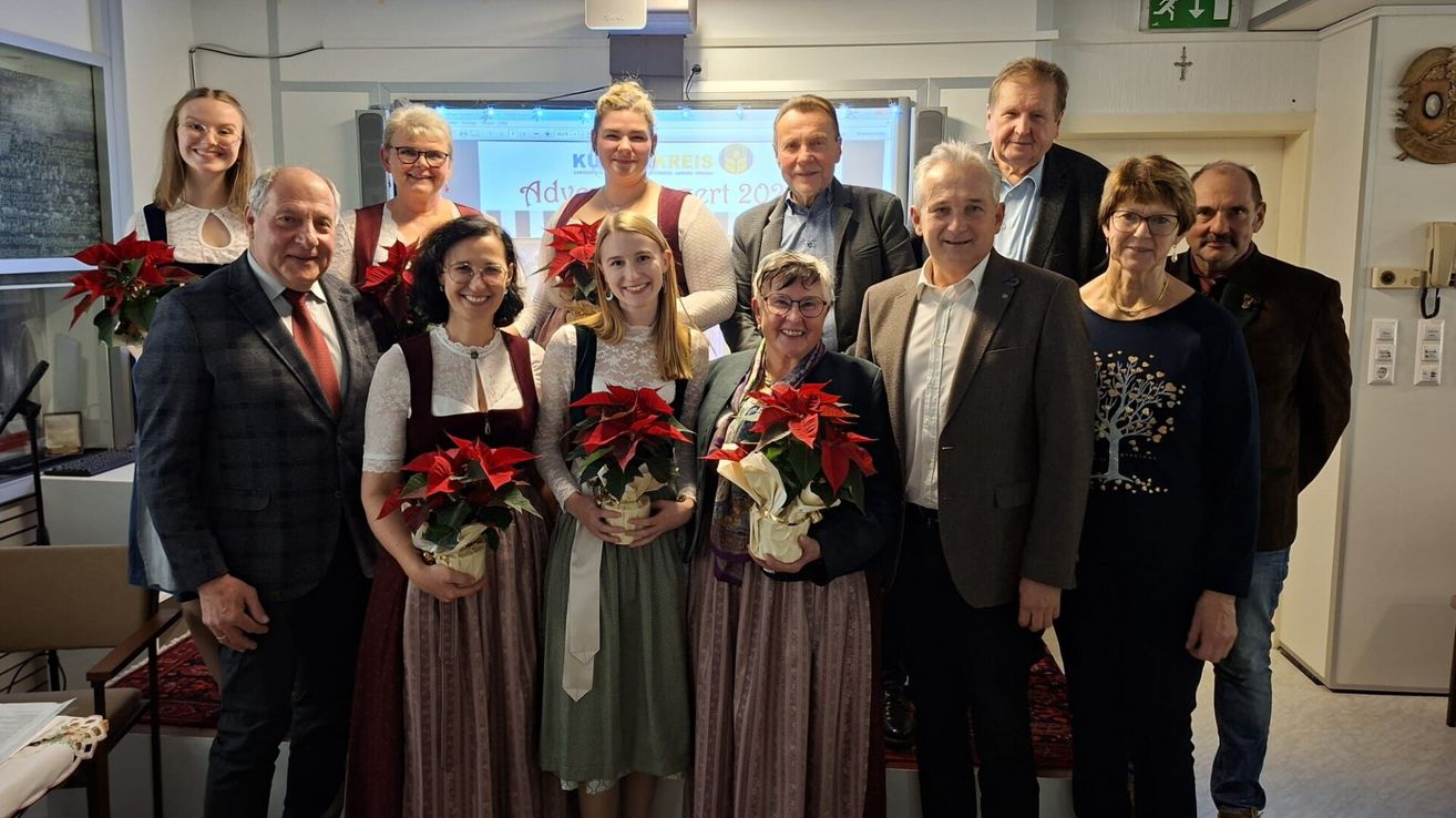 Eine Gruppe von Menschen in formeller Kleidung posiert für ein Foto, wobei Frauen in traditionellen Kleidern Blumen halten. Hinter ihnen zeigt eine Projektionsleinwand den Titel einer Veranstaltung.