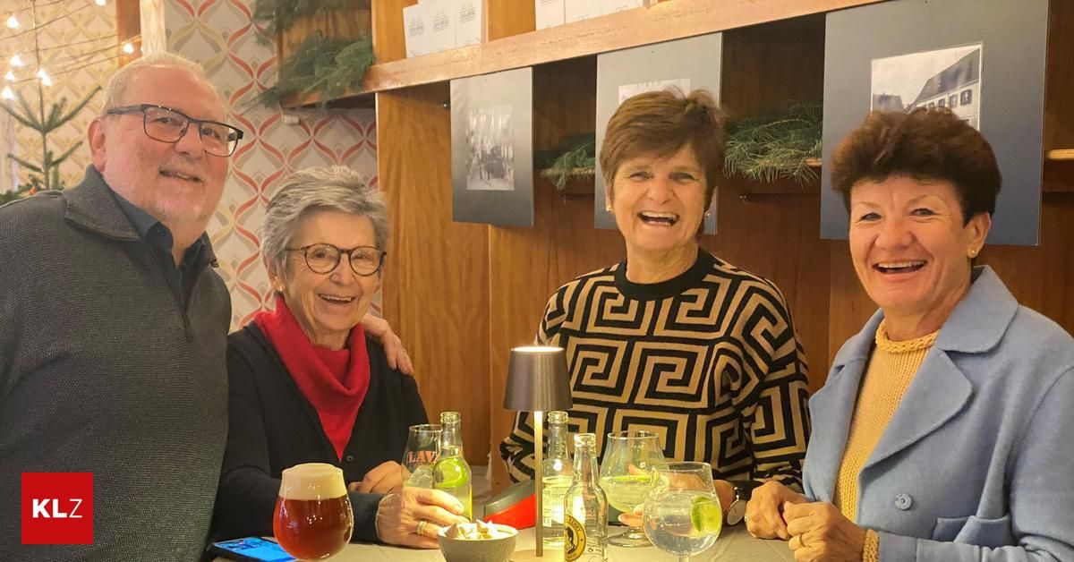 Vier Menschen sitzen an einem Tisch in einer Bar. Die Frau rechts lächelt und die Frau links hält ein Glas Bier. Dahinter befinden sich Regale mit verschiedenen Gegenständen.