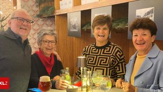 Vier Menschen sitzen an einem Tisch in einer Bar. Die Frau rechts lächelt und die Frau links hält ein Glas Bier. Dahinter befinden sich Regale mit verschiedenen Gegenständen.