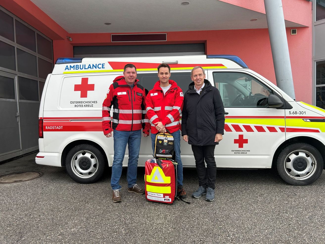 Drei Männer stehen neben einem Krankenwagen, zwei in roten Jacken, einer in Schwarz. Der Krankenwagen hat rote Kreuze und Texte, die 'Ambulanz' und 'Österreichisches Rotes Kreuz' lesen.