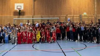 Ein Basketballteam in roten Uniformen feiert auf einem Spielfeld mit Trainern und Unterstützern, während sie Trophäen und Medaillen halten.