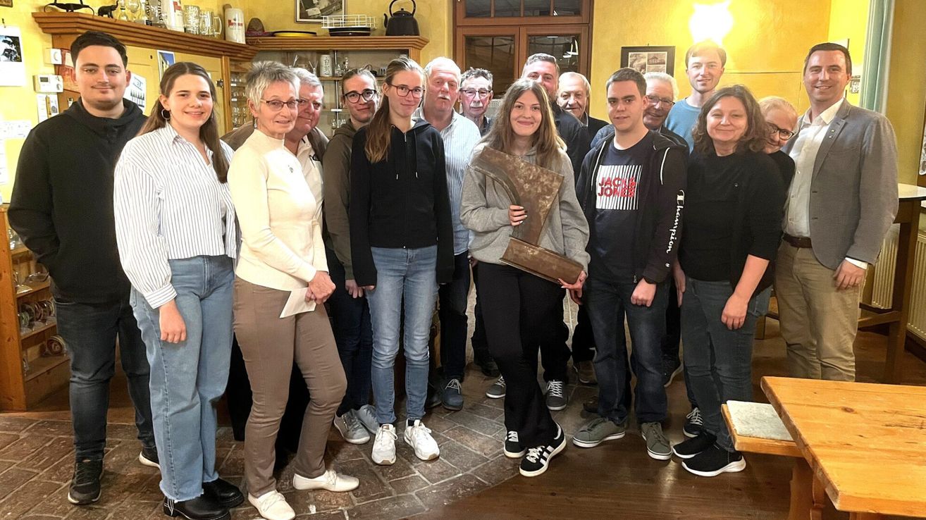 Eine Gruppe von Menschen posiert für ein Foto in einem Raum mit Holzboden. Eine Frau hält eine Trophäe und ein Mann neben ihr lächelt.
