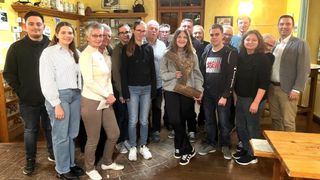 Eine Gruppe von Menschen posiert für ein Foto in einem Raum mit Holzboden. Eine Frau hält eine Trophäe und ein Mann neben ihr lächelt.