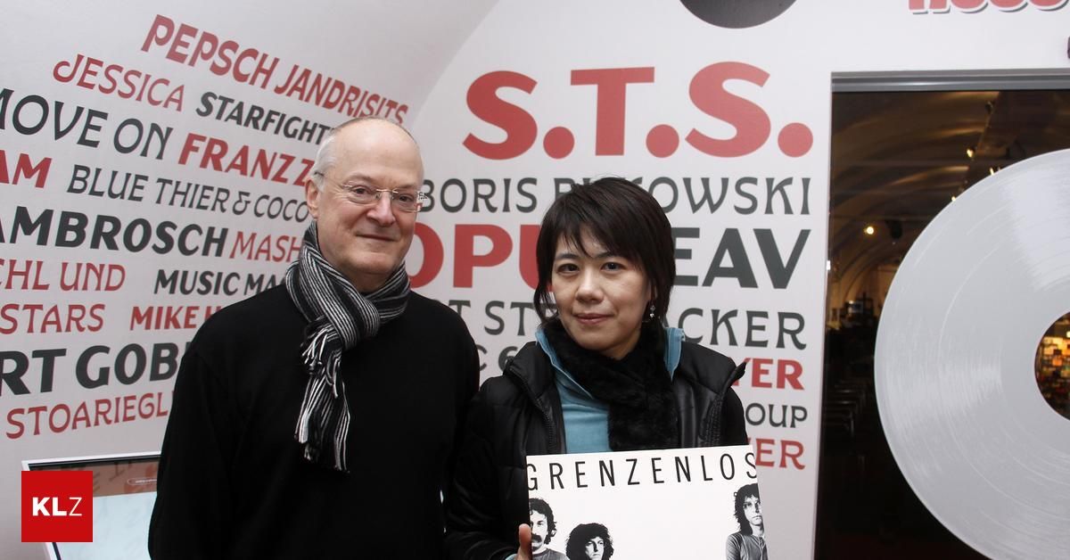 Ein Mann und eine Frau stehen nah beieinander und lächeln vor einer Wand. Der Mann trägt eine Brille und einen Schal, die Frau eine Lederjacke. Sie halten ein Buch mit dem Titel Grenzlos.