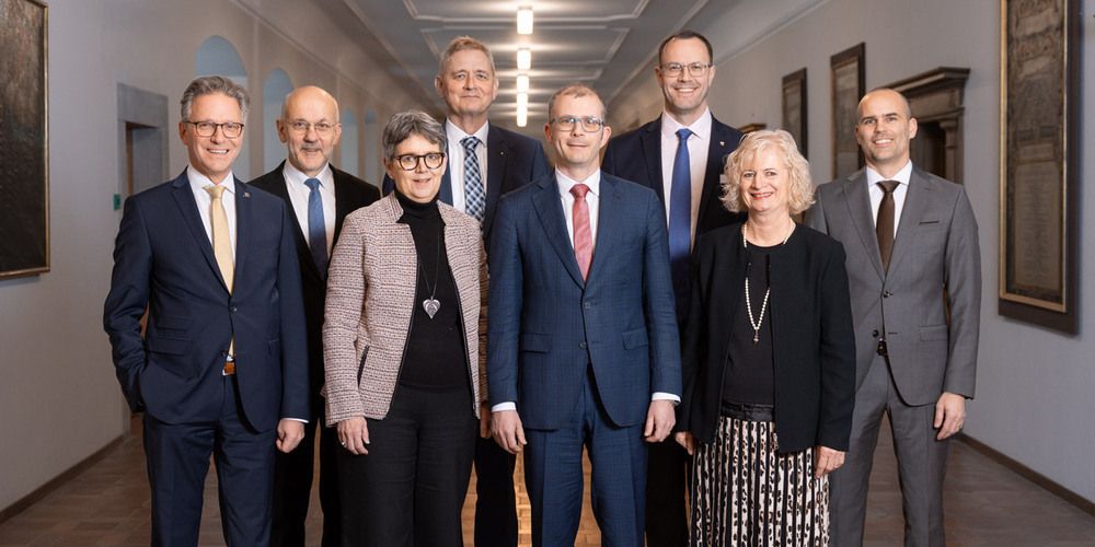 Eine Gruppe formell gekleideter Personen steht in einem Flur. Sie lächeln und scheinen für ein Foto zu posieren.