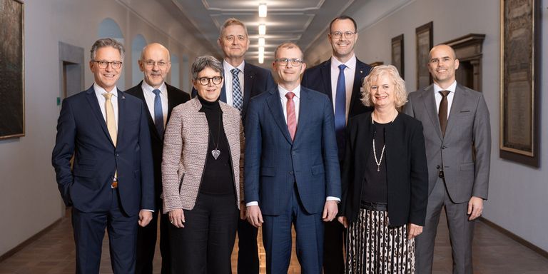 Eine Gruppe formell gekleideter Personen steht in einem Flur. Sie lächeln und scheinen für ein Foto zu posieren.