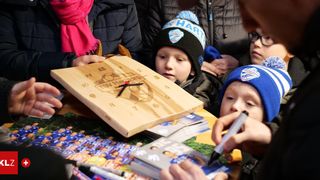 Ein junger Junge signiert eine Holzuhr mit einem Marker, umgeben von Erwachsenen und anderen Kindern, mit einem socker-thematischen Hintergrund.